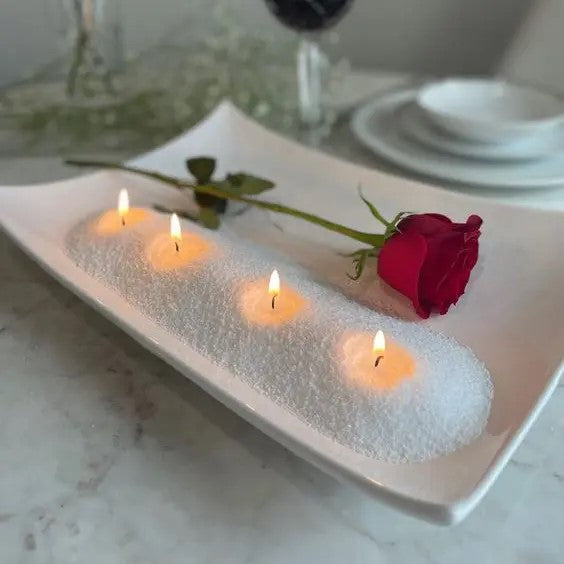 Row of small lit pearled wax candles set into a white tray of wax pearls with a single red rose, styled as a romantic dinner table centerpiece